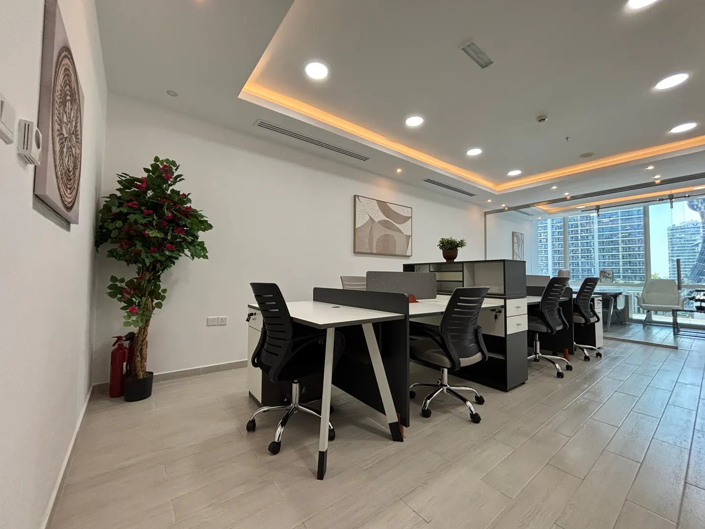 Bright office space with white walls, light wood floors, and modern desks with black chairs. A large window shows a city view. Artwork and plants add a touch of decor.