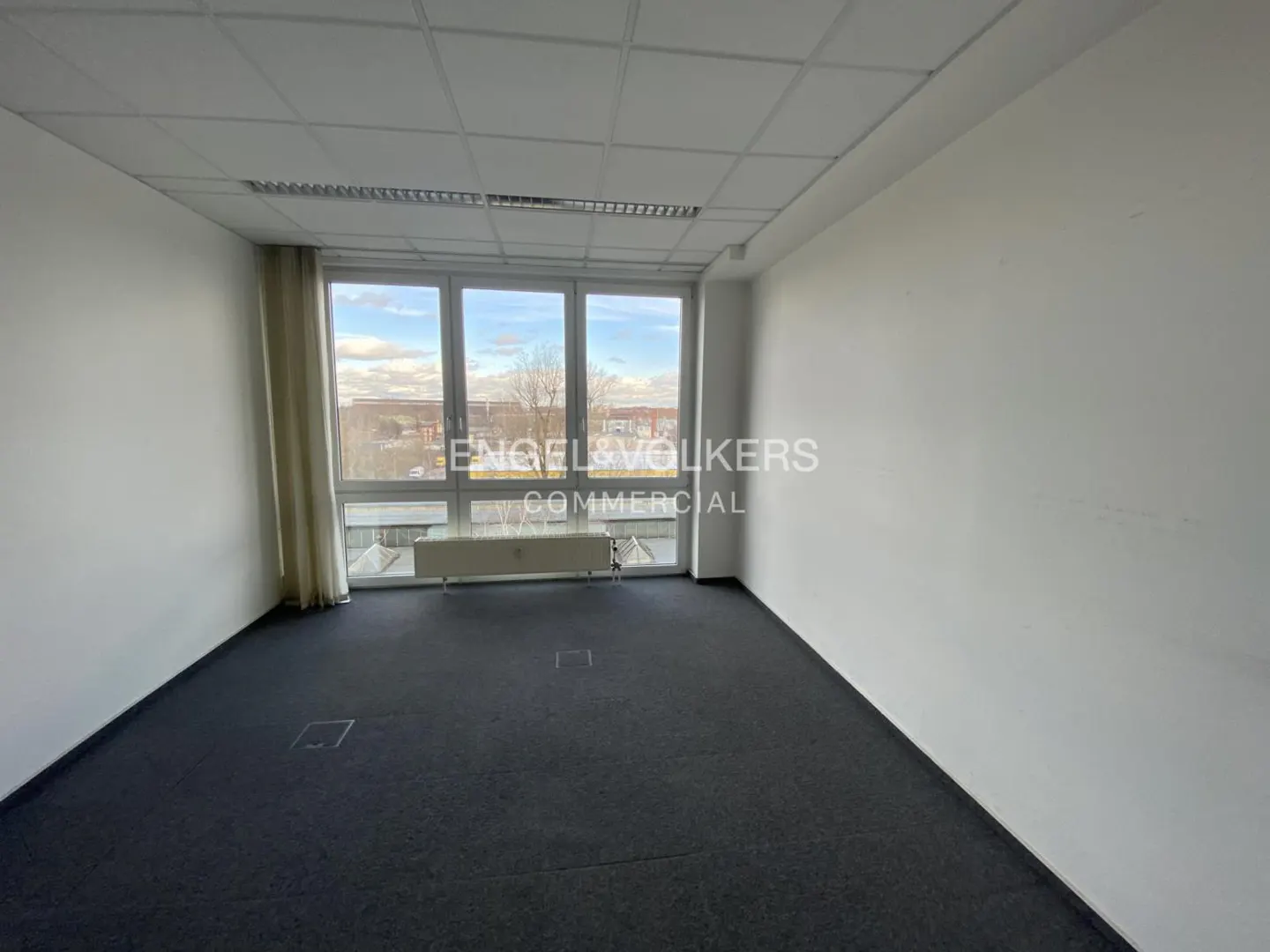 Empty office space with gray carpet, white walls, and a large window showing a cityscape. Curtains are on the left side of the window.