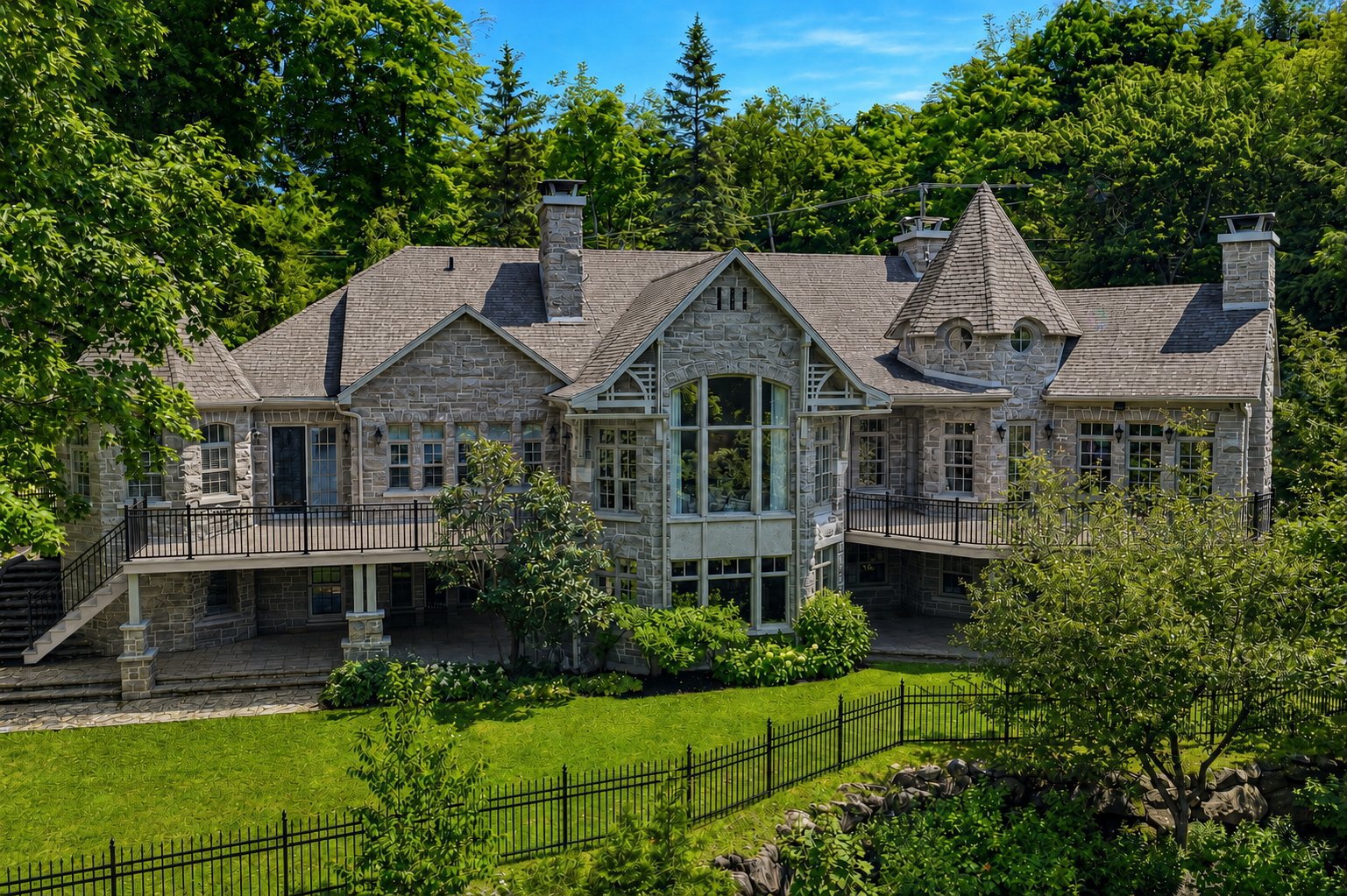 A large, grey stone mansion with a turret, multiple balconies, and a lush green backyard enclosed by a black metal fence.