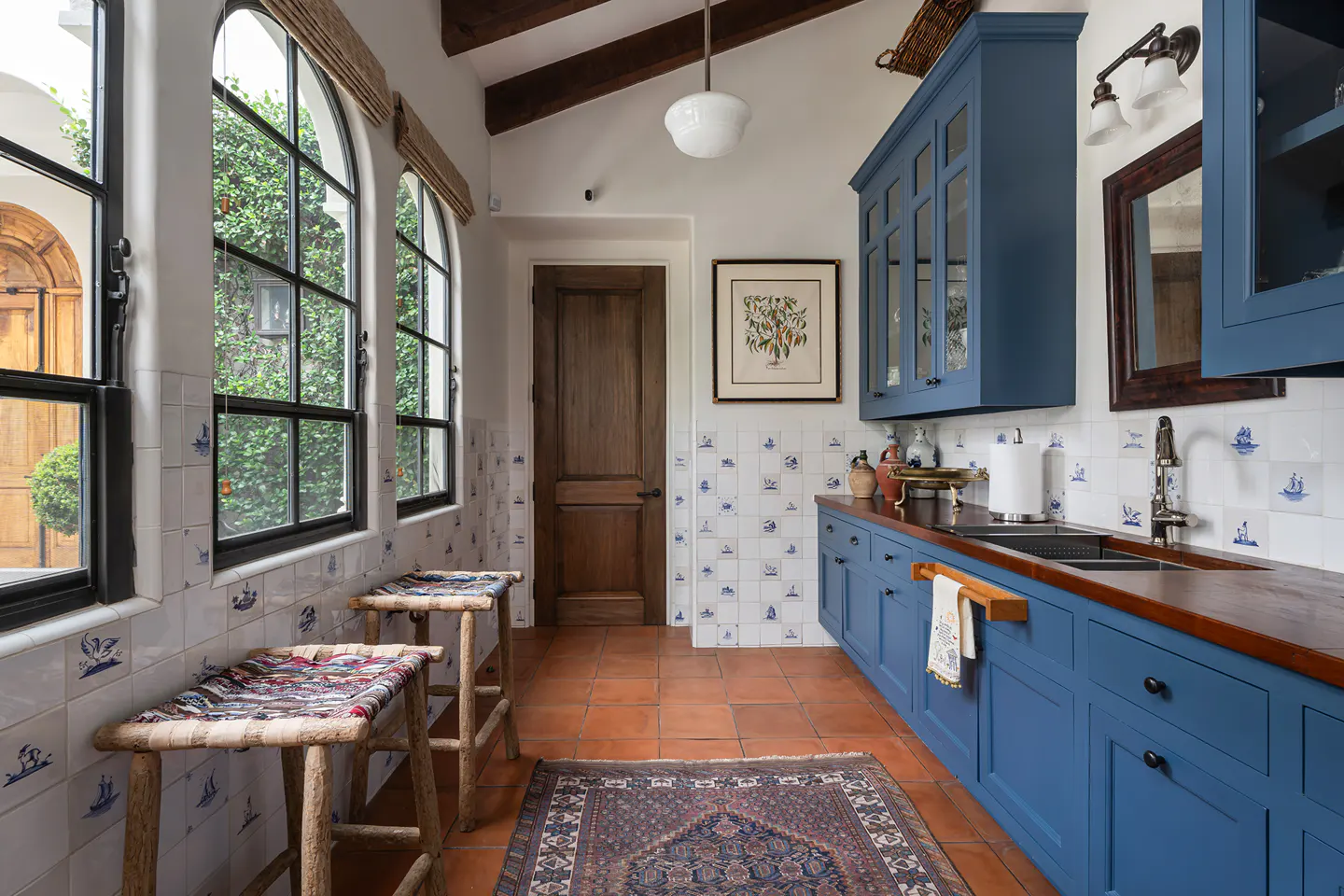 A kitchen with blue cabinets, wood countertops, and white tile backsplash with blue designs. Two stools sit near arched windows. A rug covers the terracotta floor.