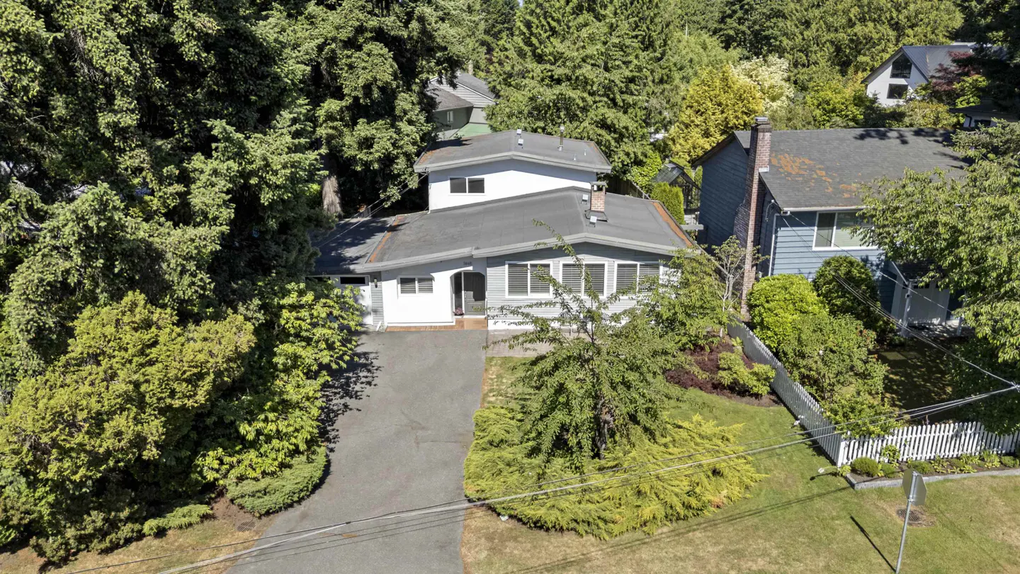Aerial view of a light gray split-level home with a paved driveway, surrounded by lush green trees and a white fence.