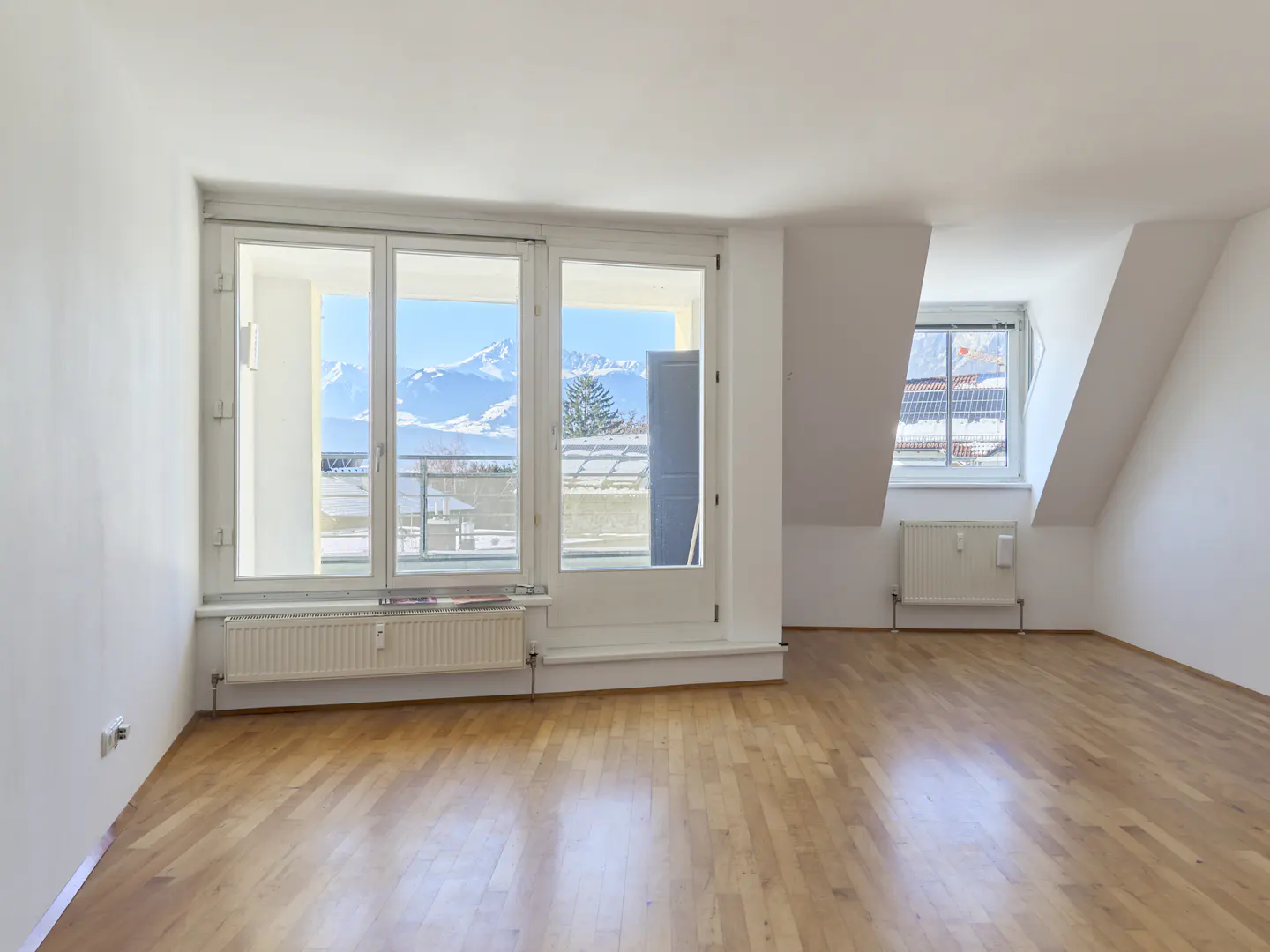 Bright, empty room with hardwood floors, white walls, and large windows with mountain views. Radiators are under the windows.
