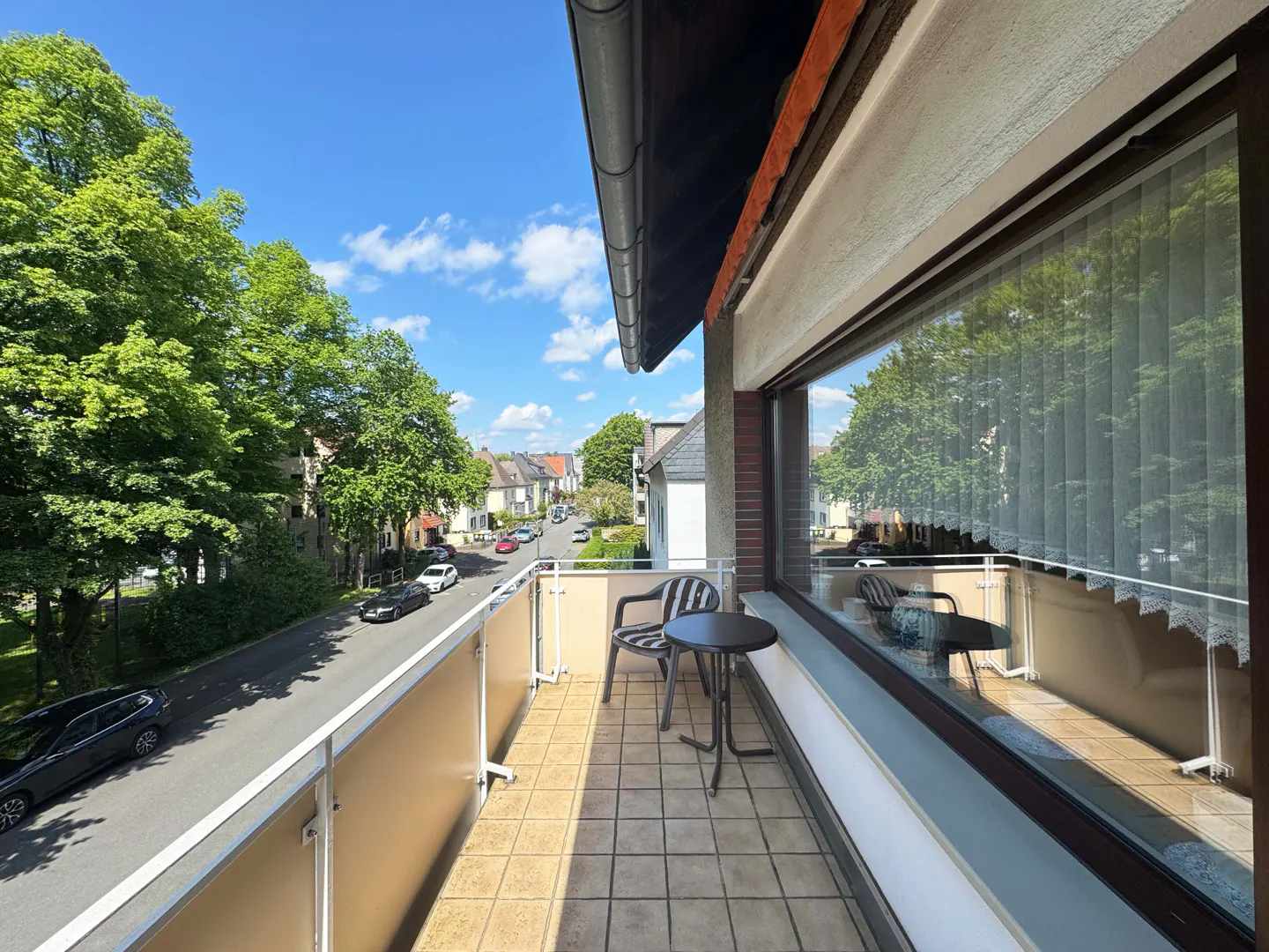 Balcony view with a chair and table overlooking a street with trees and parked cars on a sunny day.