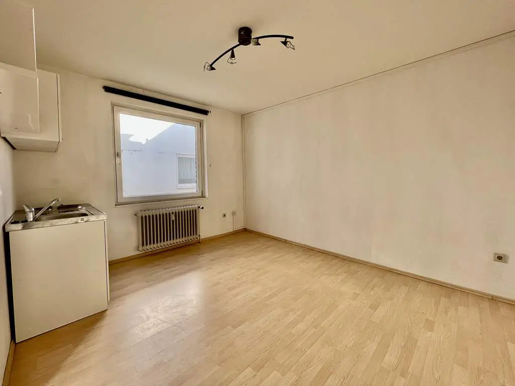 Empty room with light wood floors, white walls, and a window. A small kitchenette is in the corner. A radiator sits under the window.