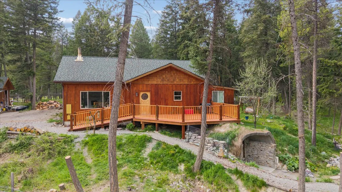 A wood-sided house with a green roof and a wooden deck, surrounded by tall trees. A stone-lined tunnel entrance is visible on the hillside.