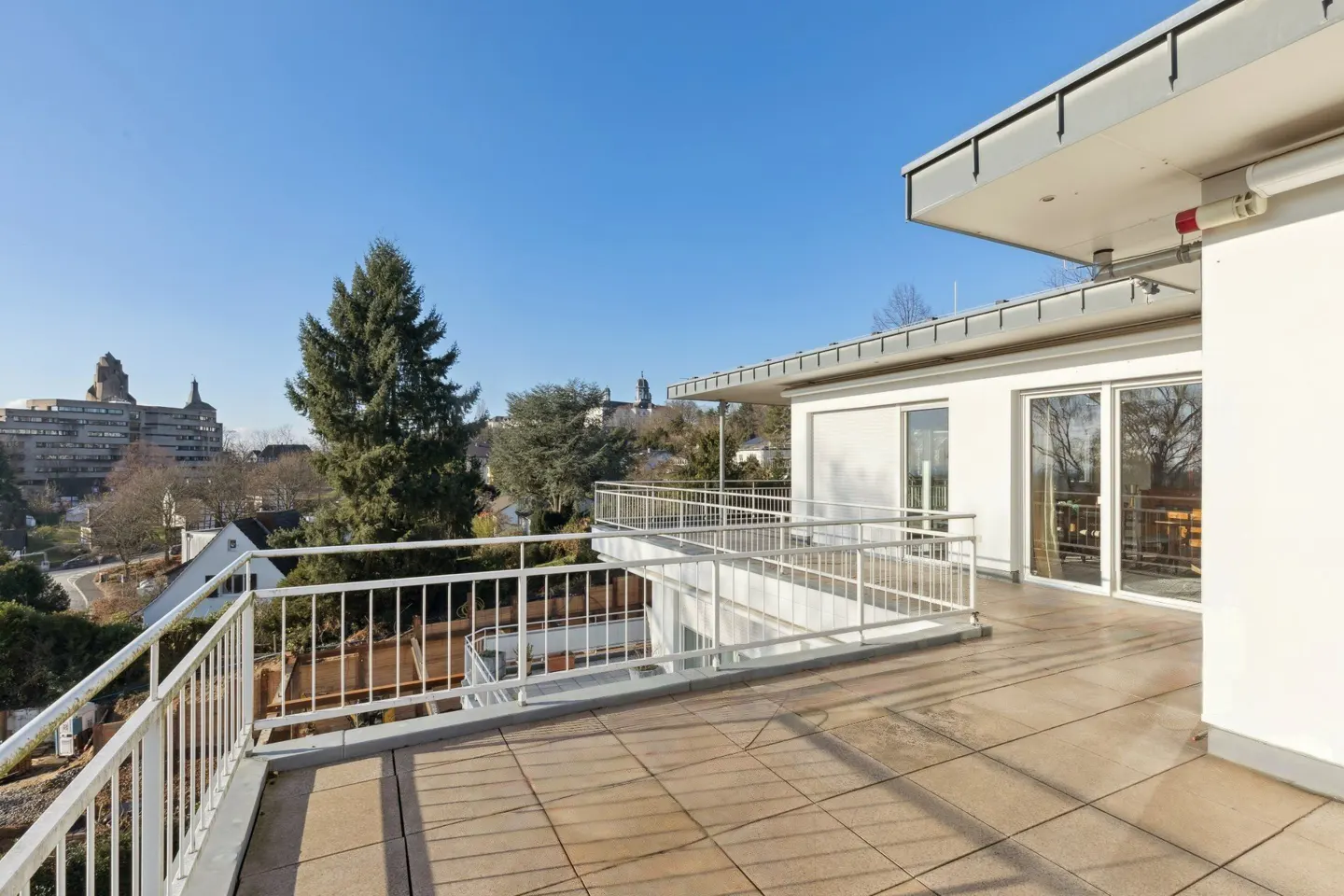 Wide balcony with white railings and tile flooring, overlooking a cityscape with trees and buildings under a clear blue sky.