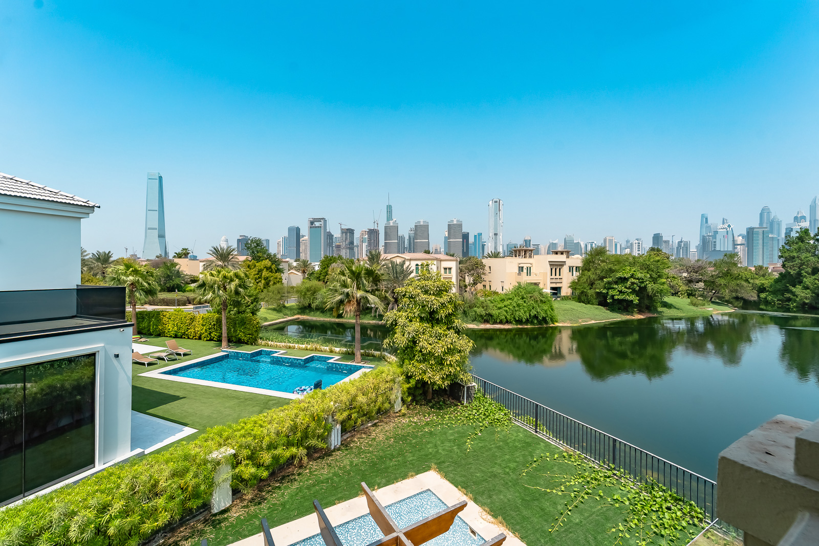 Luxury villa backyard with a private pool, green lawn, and lake, overlooking a distant Dubai city skyline under a blue sky. [AI-generated text]