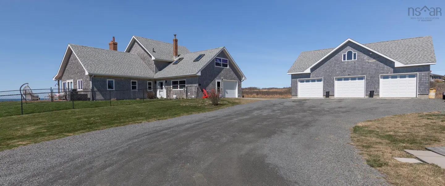 Exterior view of a gray shingle house with a matching three-car garage on a gravel driveway under a clear blue sky.