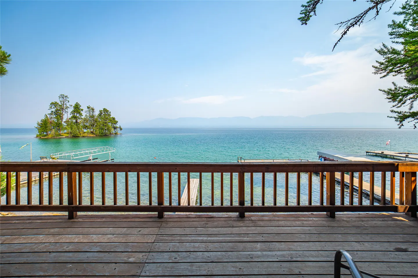 View from a wooden deck with a brown railing overlooking a turquoise lake with docks and a small, tree-covered island under a blue sky.