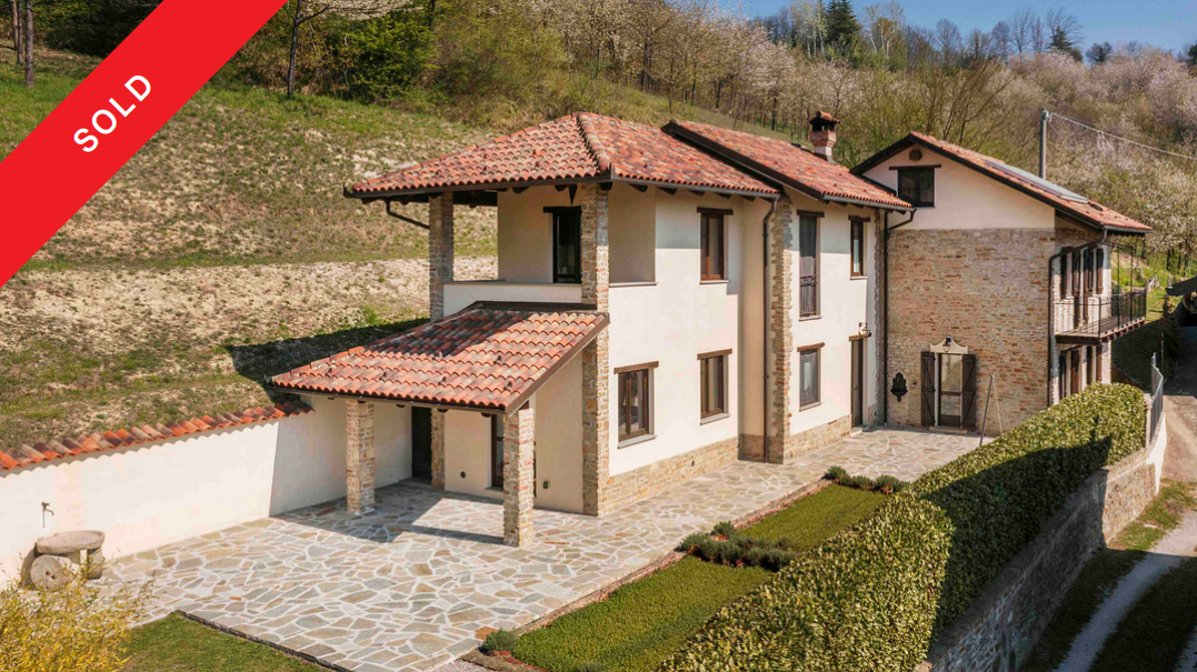 A two-story stone and stucco house with a terracotta tile roof, stone patio, and manicured hedges, marked as sold. [AI-generated text]