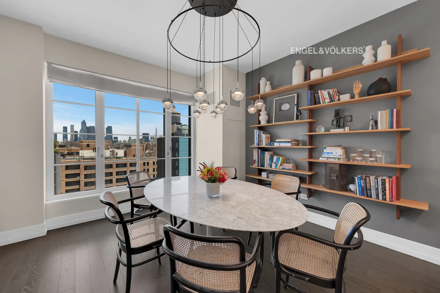 A dining room with a marble table, black chairs, and a city view. A modern chandelier hangs above the table. A bookshelf is on the right wall.