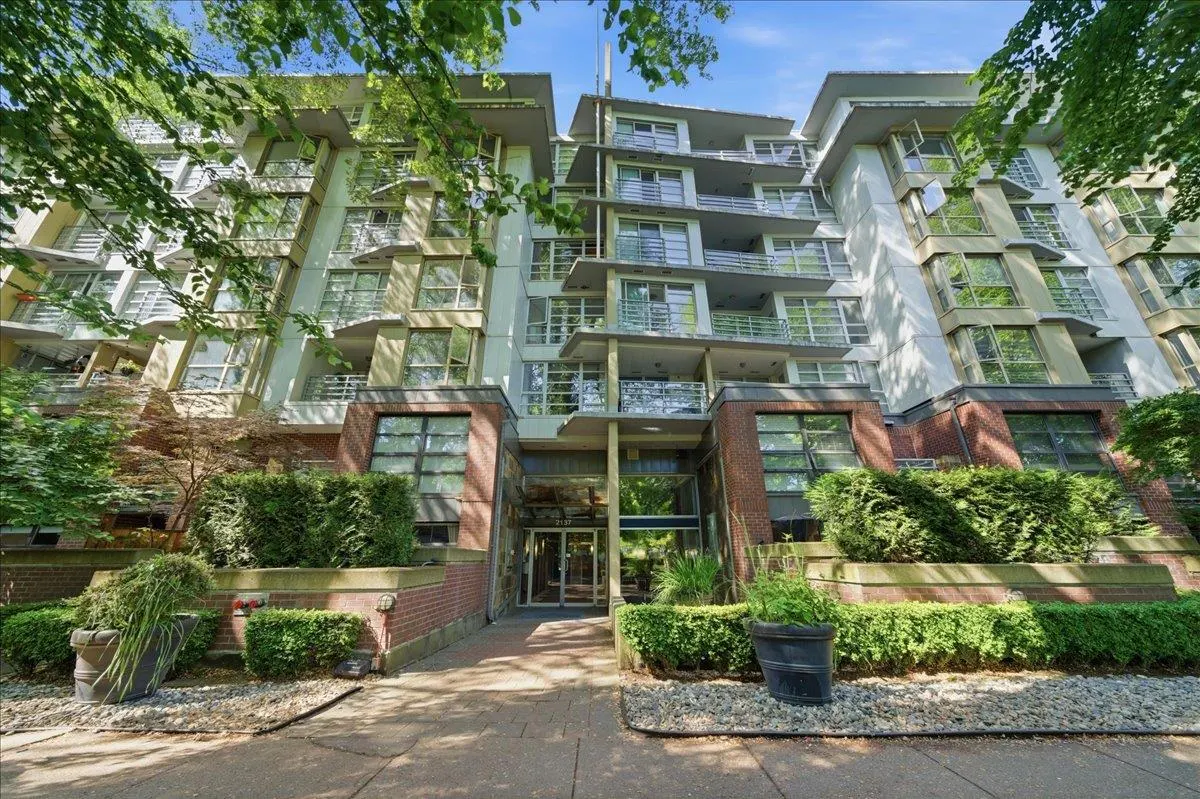Low-angle view of a modern apartment building with brick base, white upper levels, balconies, and lush green landscaping.