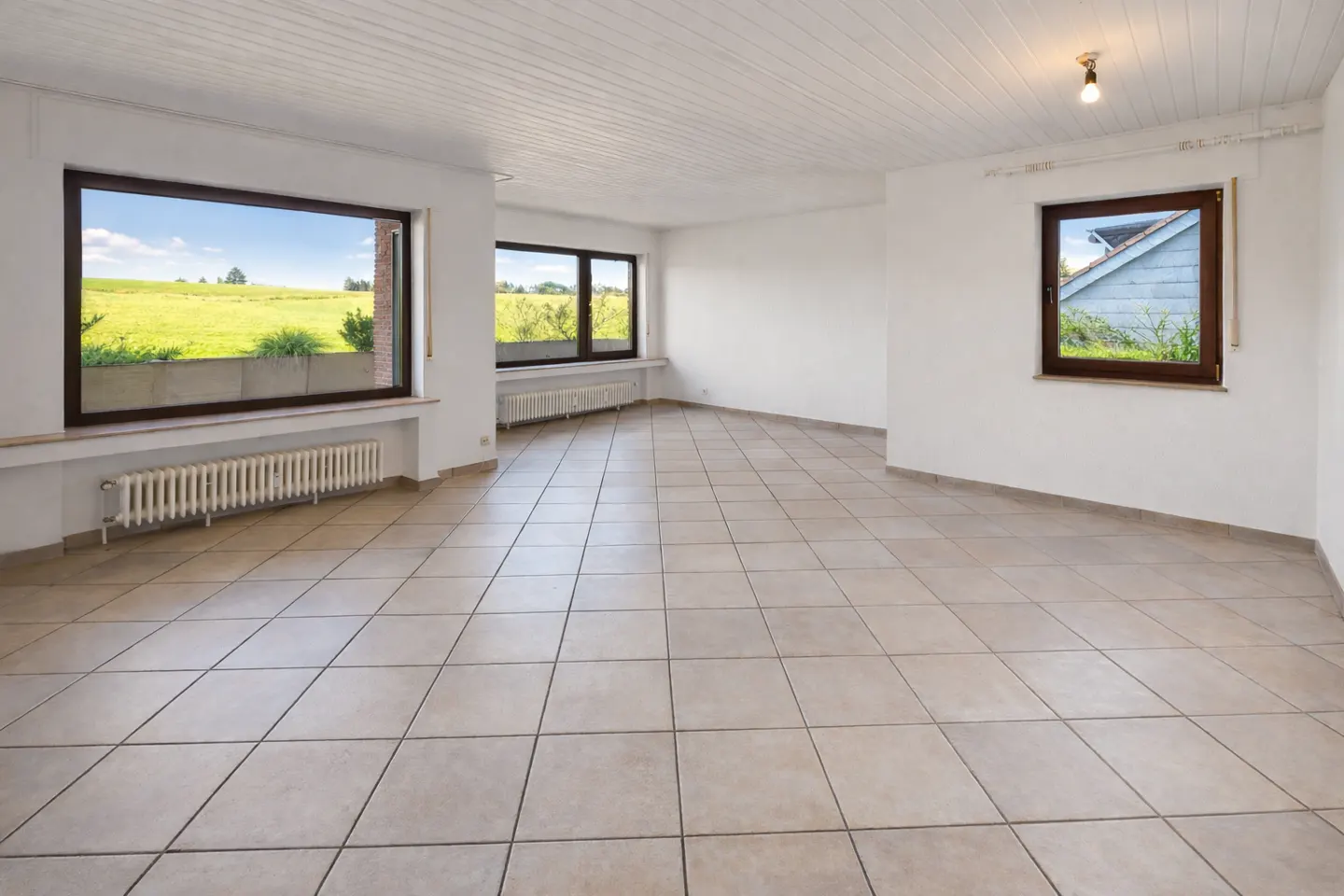 Empty room with beige tile floor, white walls, and wood-framed windows showing green fields. A white radiator sits under the largest window.