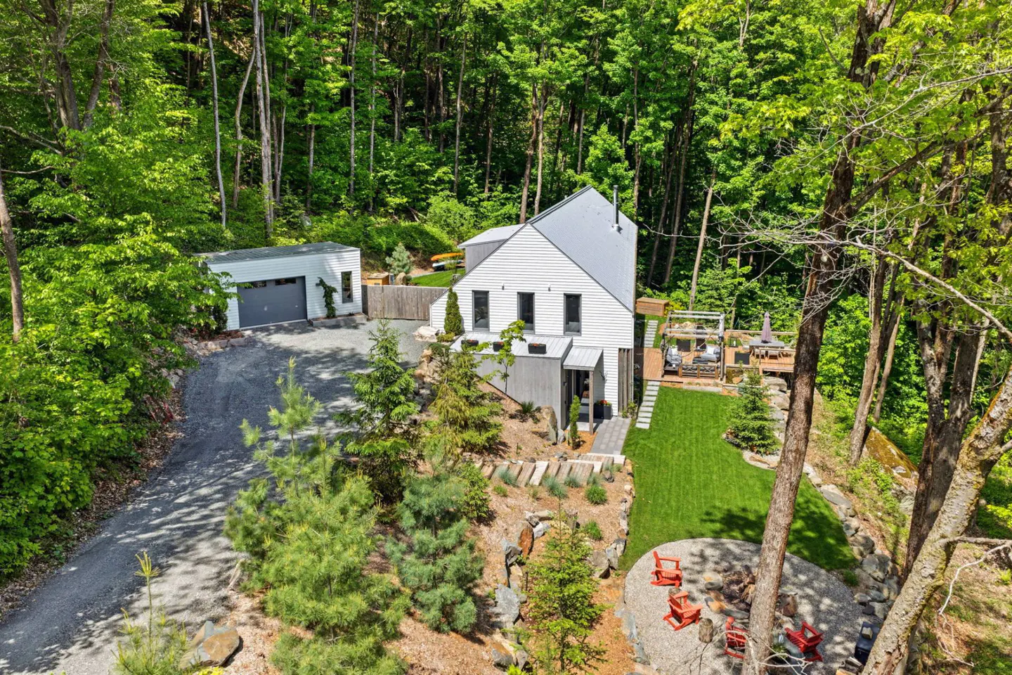 Aerial view of a white modern home with a detached garage, lush forest, green lawn, and a fire pit with red chairs.