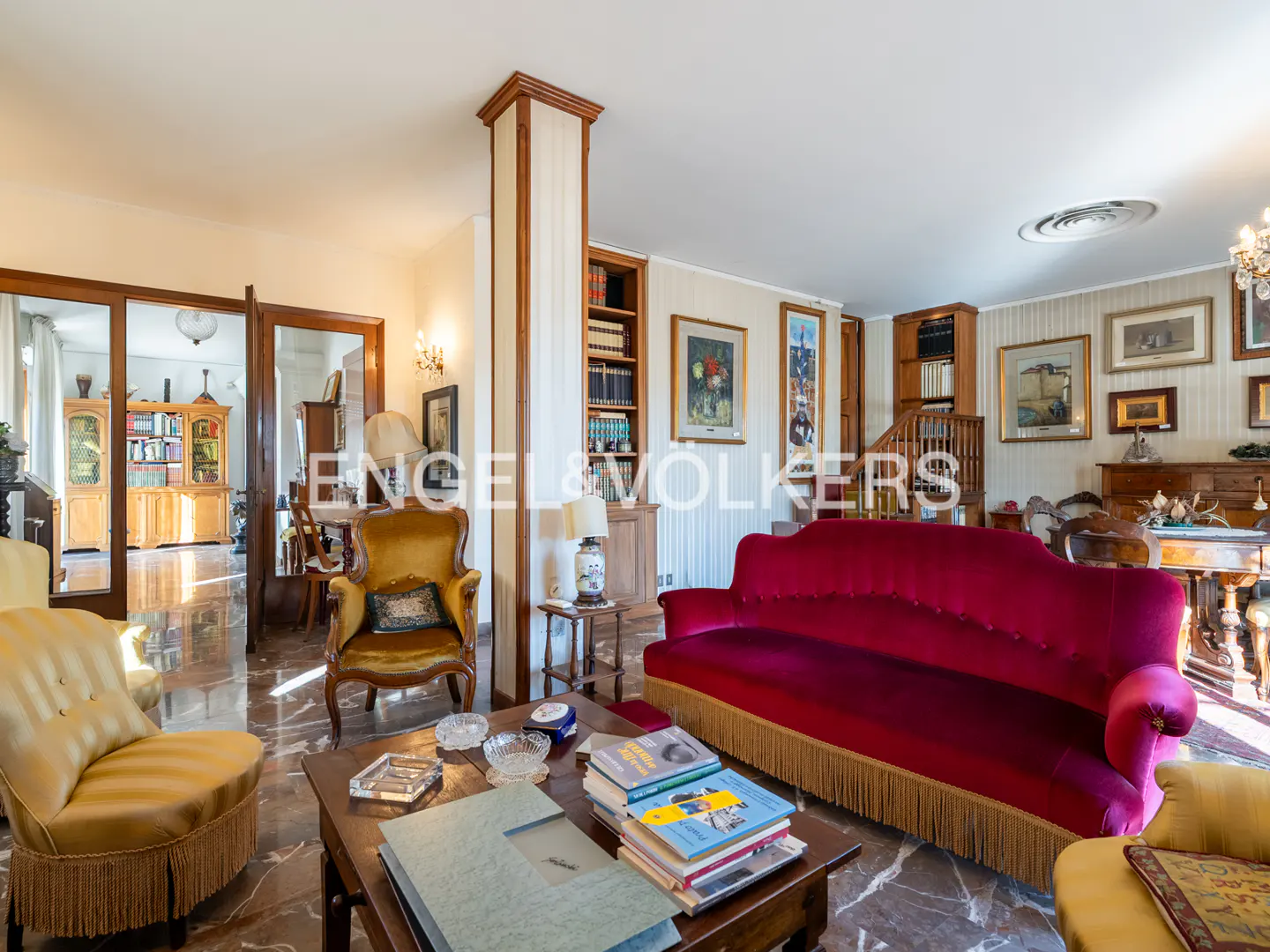 A living room with marble floors, yellow armchairs, and a red velvet sofa. Books are stacked on a dark wood coffee table.