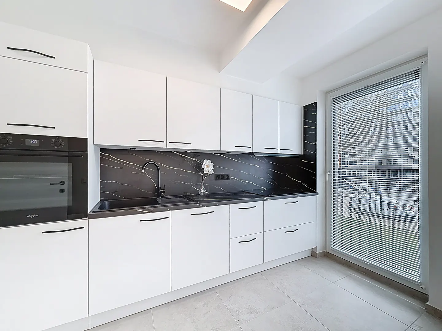 Bright kitchen with white cabinets, black marble backsplash, and a black sink. A window with blinds overlooks a building.