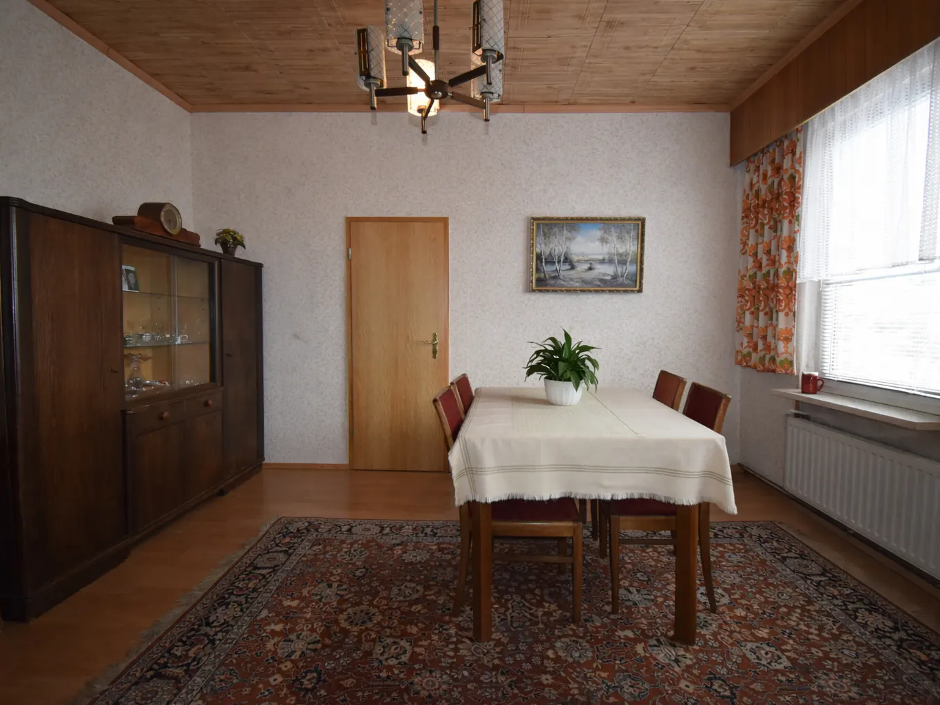 A dining room with a wooden table covered by a white tablecloth, surrounded by red chairs. A brown cabinet stands against the wall. A patterned rug covers the floor.
