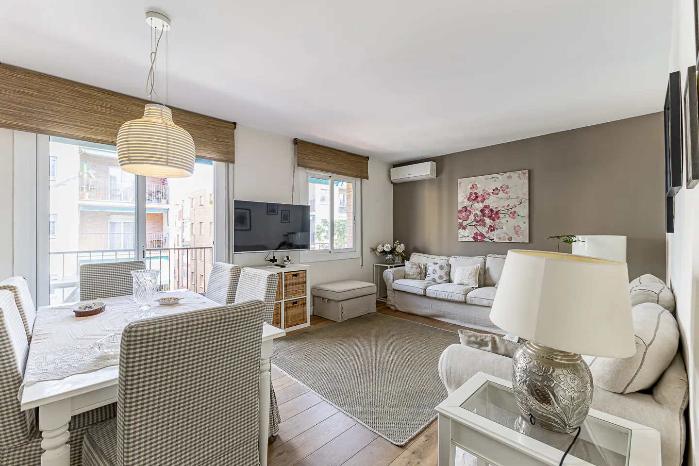 Bright living room with dining area. White table and chairs, beige sofa, and a painting with pink flowers on a gray wall.