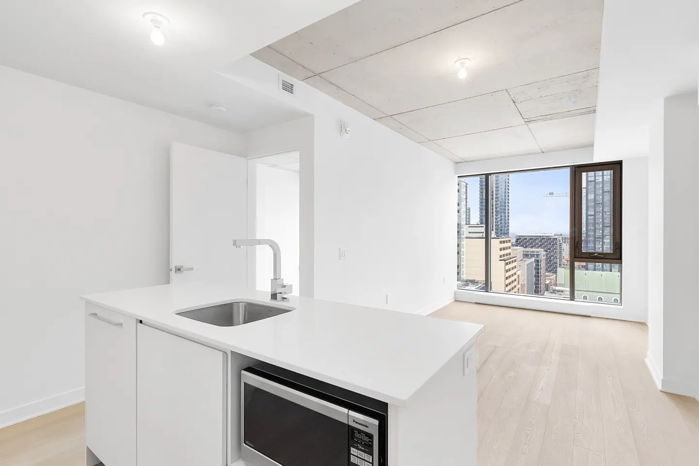 Modern kitchen island with sink and microwave in a bright apartment with concrete ceiling and city view through windows.