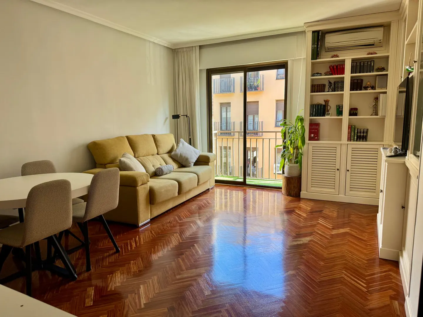 Bright living room with herringbone wood floors, a yellow sofa, dining table, and a white bookcase by a sliding glass door.