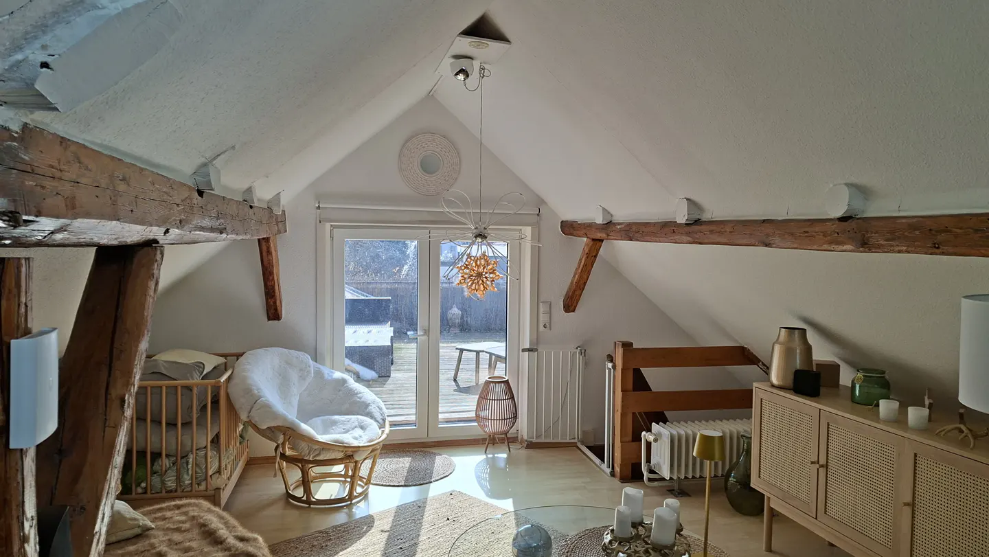 Attic room with exposed beams, white walls, and a white papasan chair. A glass door leads to a backyard. A rattan cabinet sits to the right.