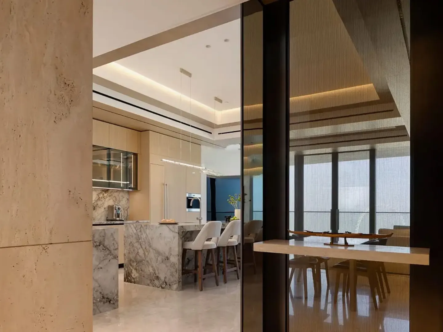 Modern kitchen with a marble island, white bar stools, and light wood cabinetry, viewed through a dark glass partition.