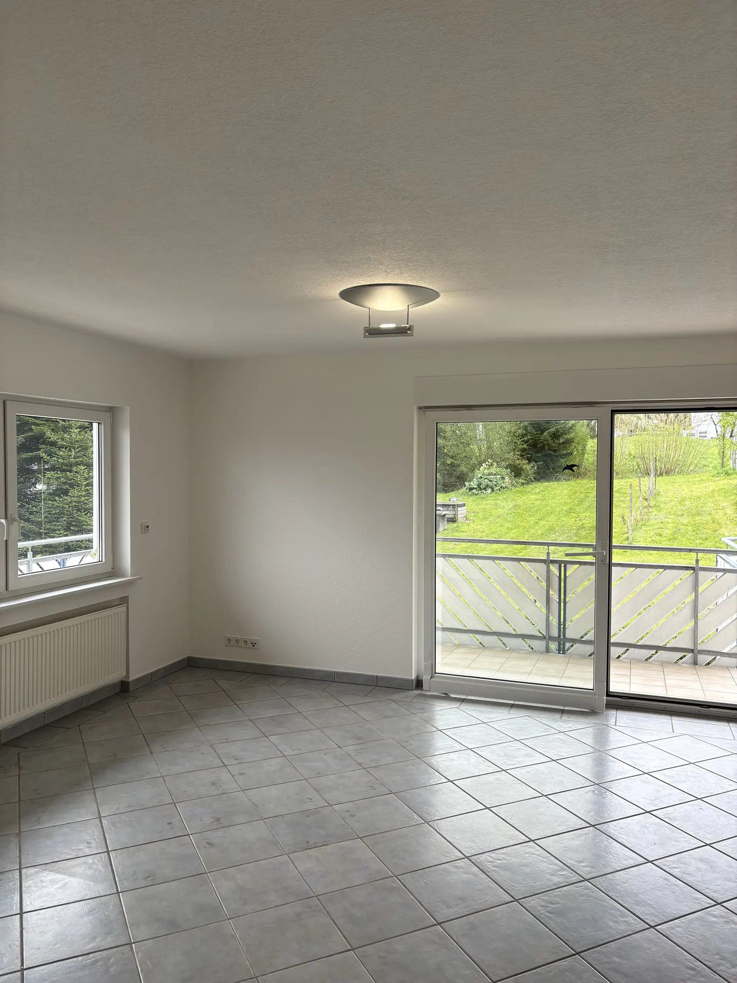 Bright, empty room with gray tile floor, white walls, and a modern ceiling light. Sliding glass doors lead to a balcony with a green lawn view. A window with a radiator is on the left.
