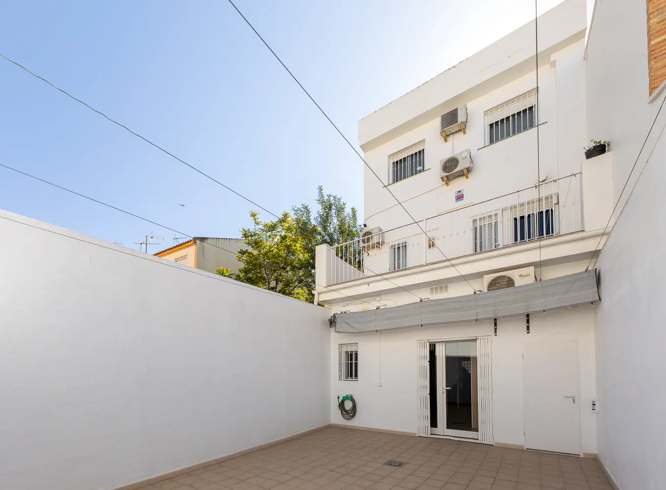 A white walled patio with a tiled floor, a white building with windows, and a blue sky.