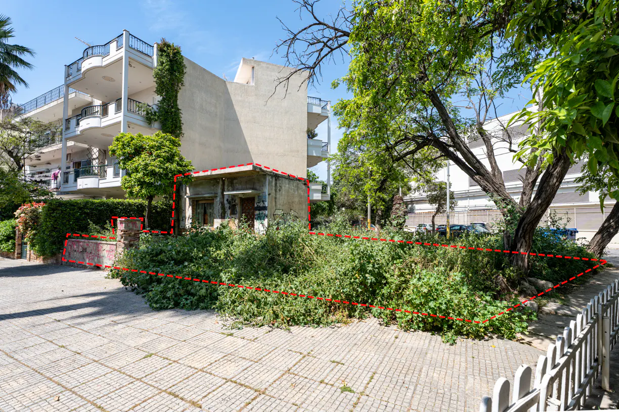 Real estate image shows a vacant lot with overgrown greenery, a small building, and a multi-story apartment building in the background.