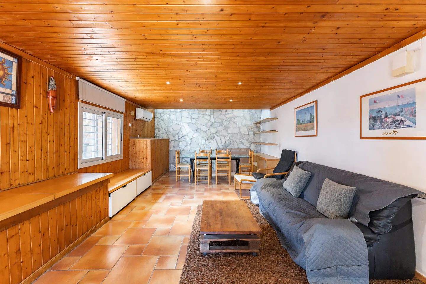 Living room with wood-paneled walls and ceiling, tile floor, gray sofa, and dining table with chairs.