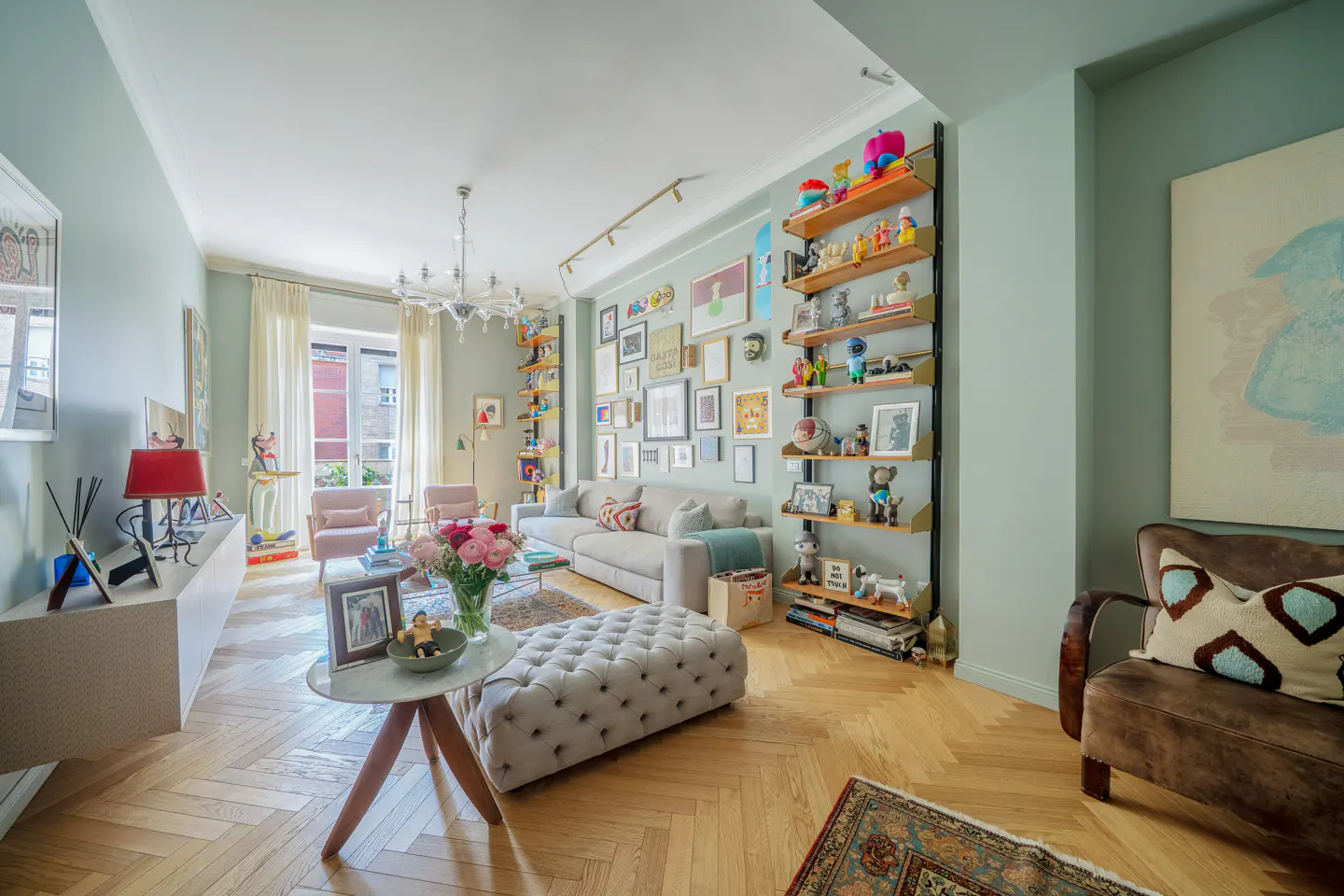 Bright living room with light green walls, herringbone wood floors, and eclectic decor. Shelves display toys and art. A tufted ottoman sits near a marble table.