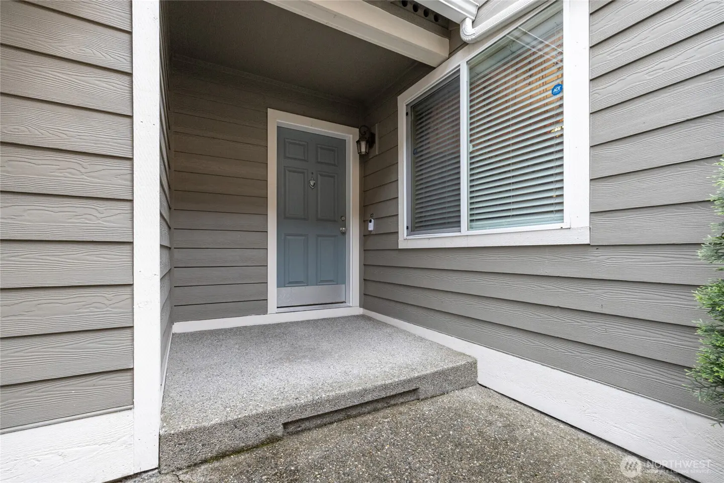 Exterior view of a home's entrance with gray siding, a blue door, and a concrete porch. A window with blinds is to the right of the door.