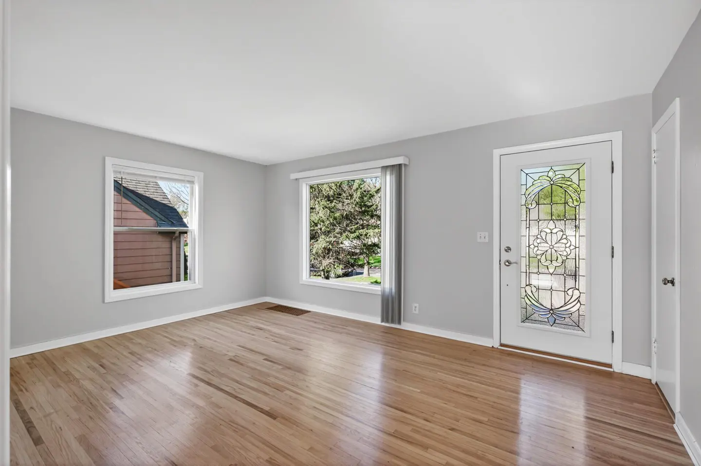 Bright, empty living room with hardwood floors, gray walls, and white trim. Windows and a decorative glass door let in natural light.