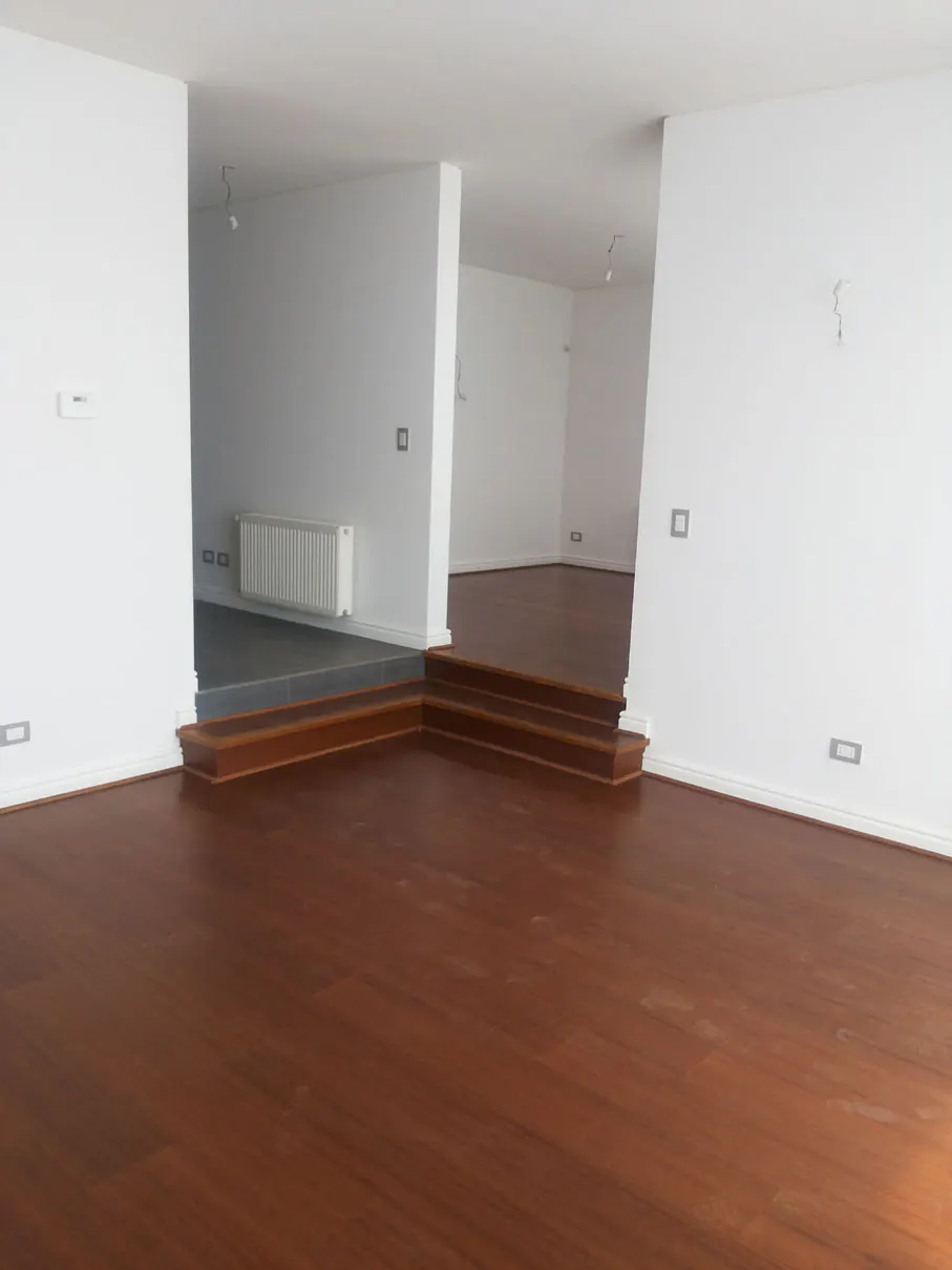 A bright, empty room with dark wood floors and white walls. Steps lead to a raised platform with a radiator.