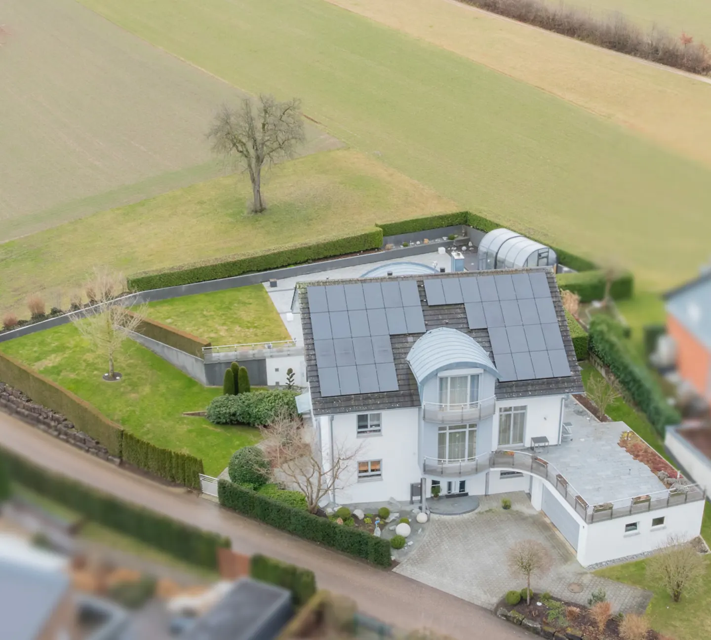 Aerial view of a modern white house with solar panels, a pool, and green lawn surrounded by fields.