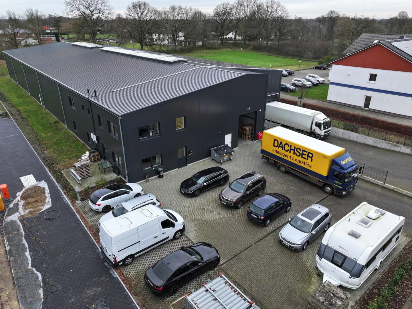 Aerial view of a dark gray warehouse with a "DACHSER" truck, cars, and a camper parked outside.