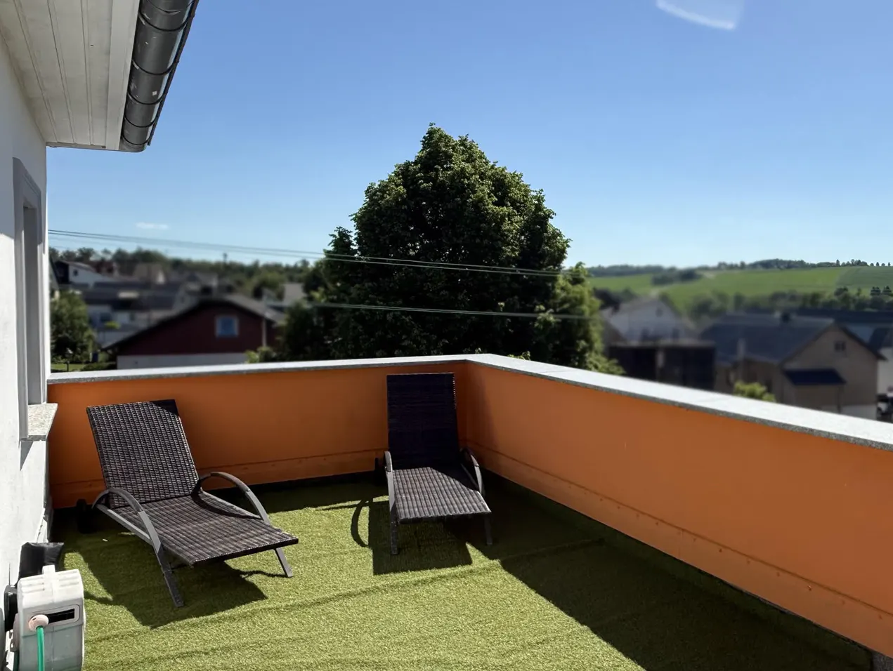 Balcony with two dark wicker lounge chairs on green turf. Orange wall, and a view of houses and trees in the background.