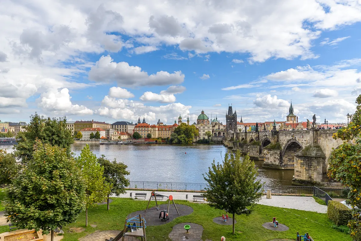 View of Prague's Charles Bridge over the Vltava River from a park with playground equipment. Blue sky with clouds.