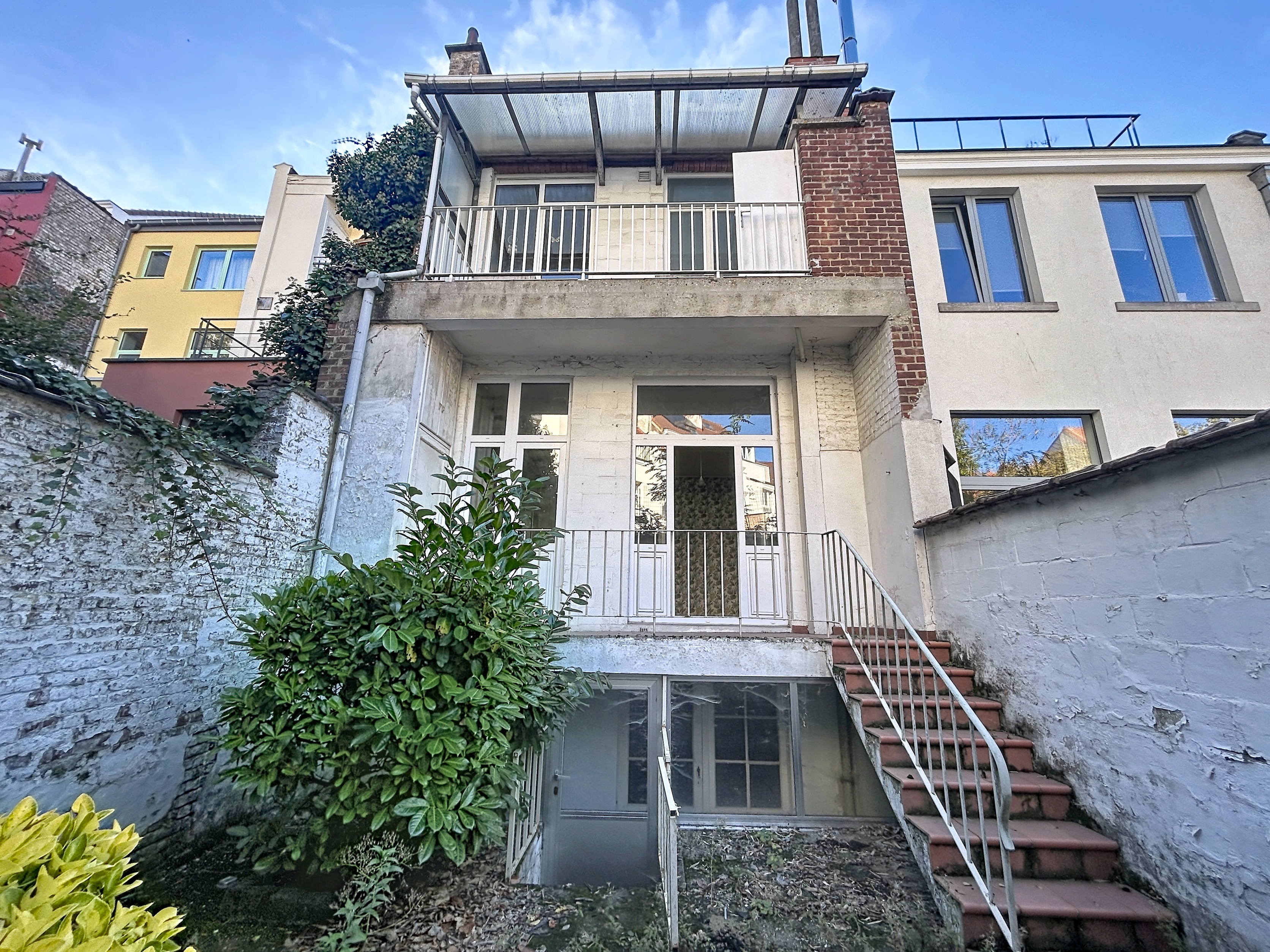 Rear view of a multi-story townhouse with a balcony, a basement entrance, and outdoor stairs leading to a small garden area. [AI-generated text]