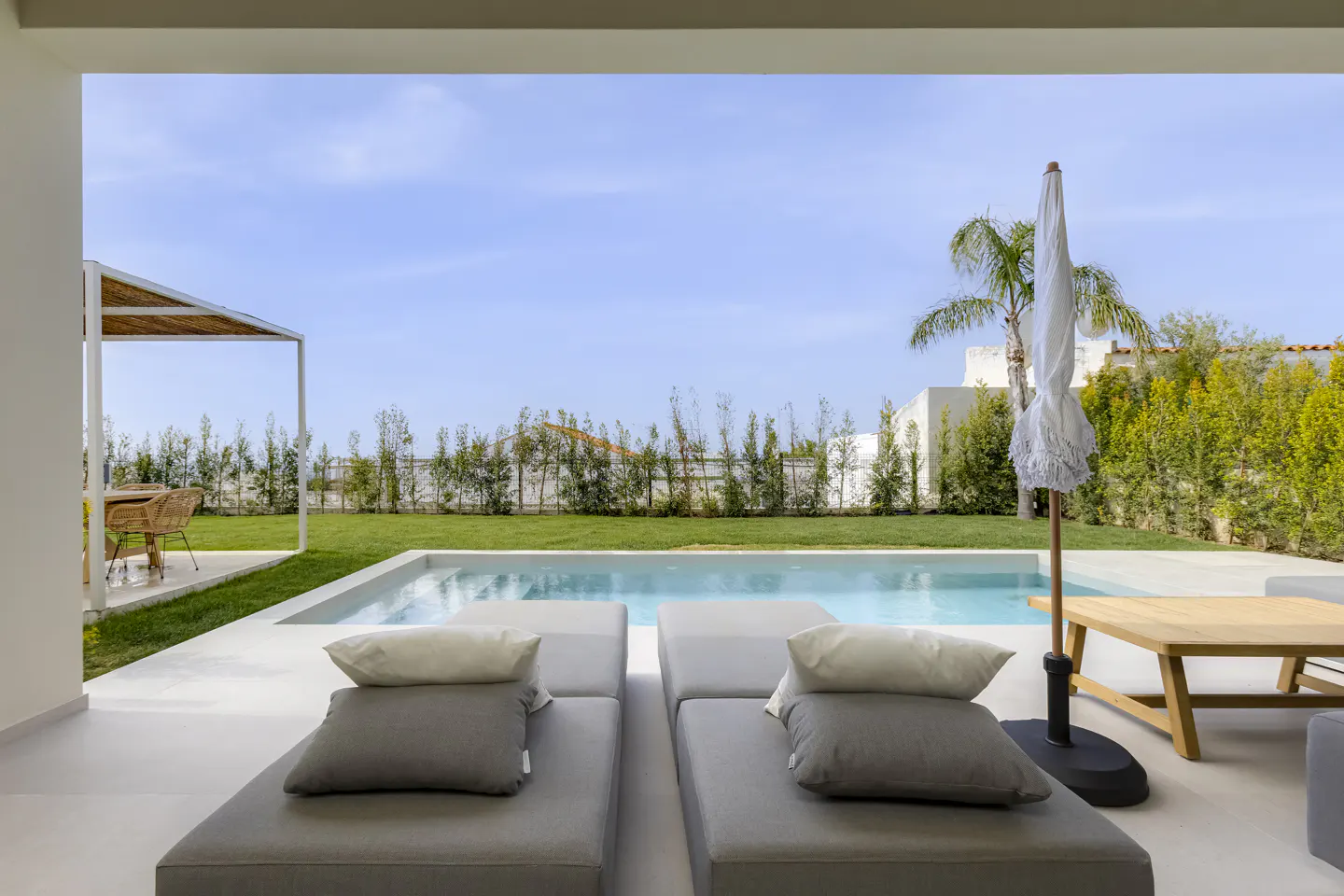 Luxury patio with gray lounge chairs, white pillows, and a pool. Green lawn, blue sky, and a white umbrella in the background.