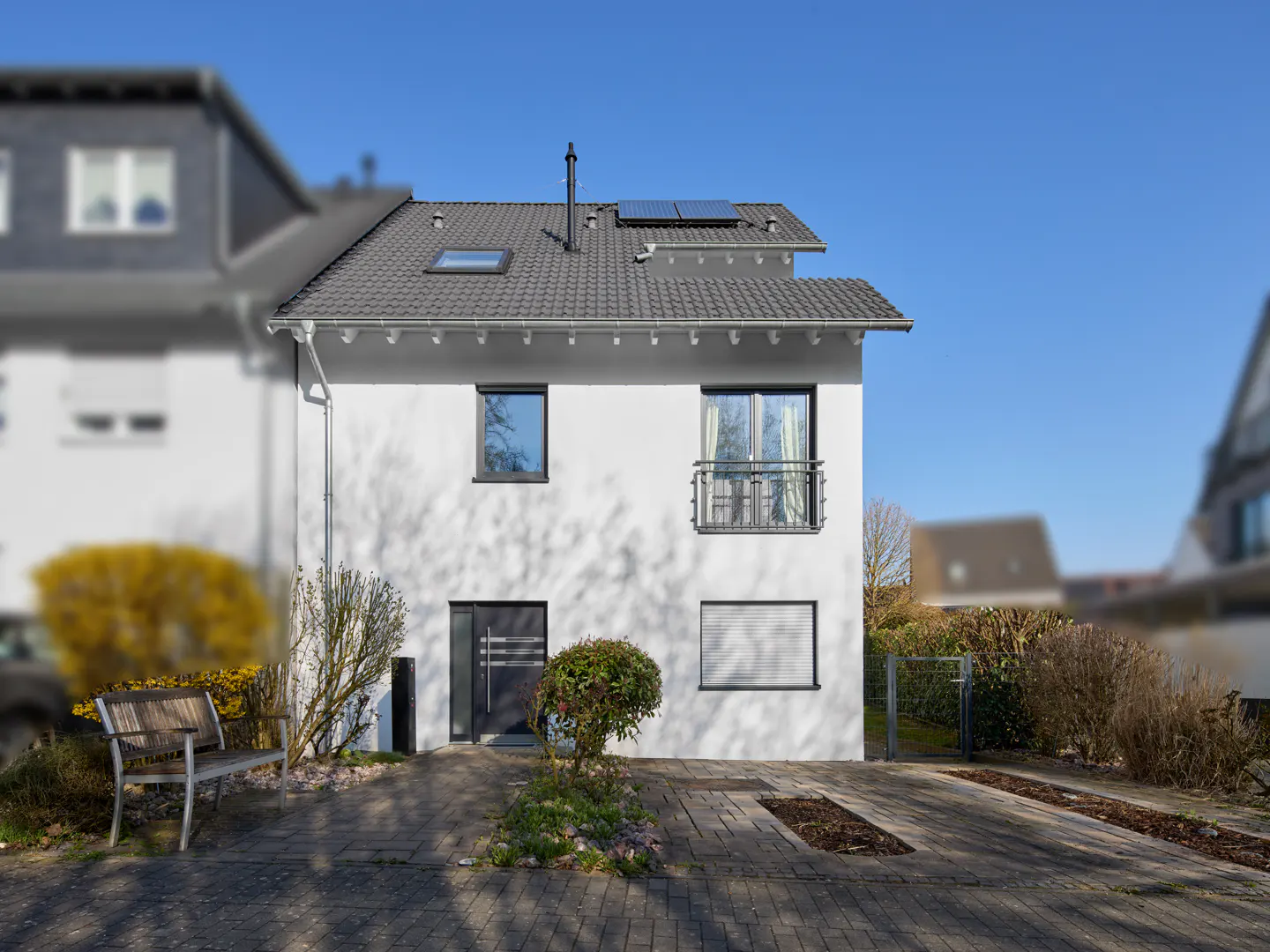 Modern two-story white house with gray roof, solar panels, and a small balcony. Paved driveway and garden in front.