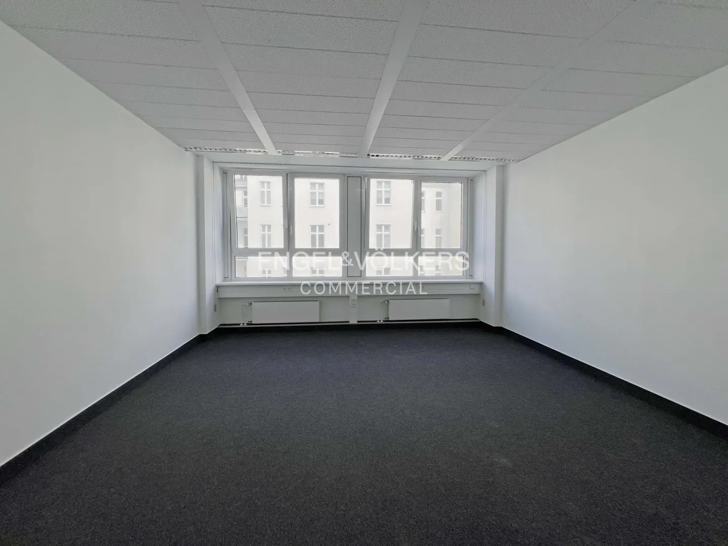 Empty office space with white walls, a dark gray carpet, and a large window with the Engel & Volkers Commercial logo.