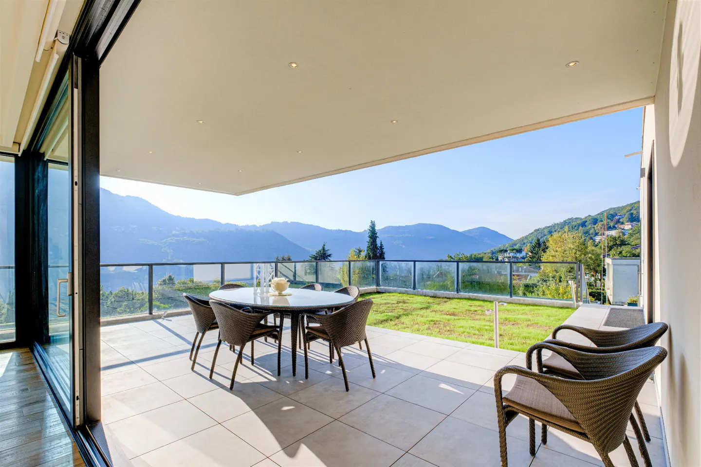 Outdoor patio with a round table, six chairs, and a mountain view. The patio has a glass railing and a tiled floor.
