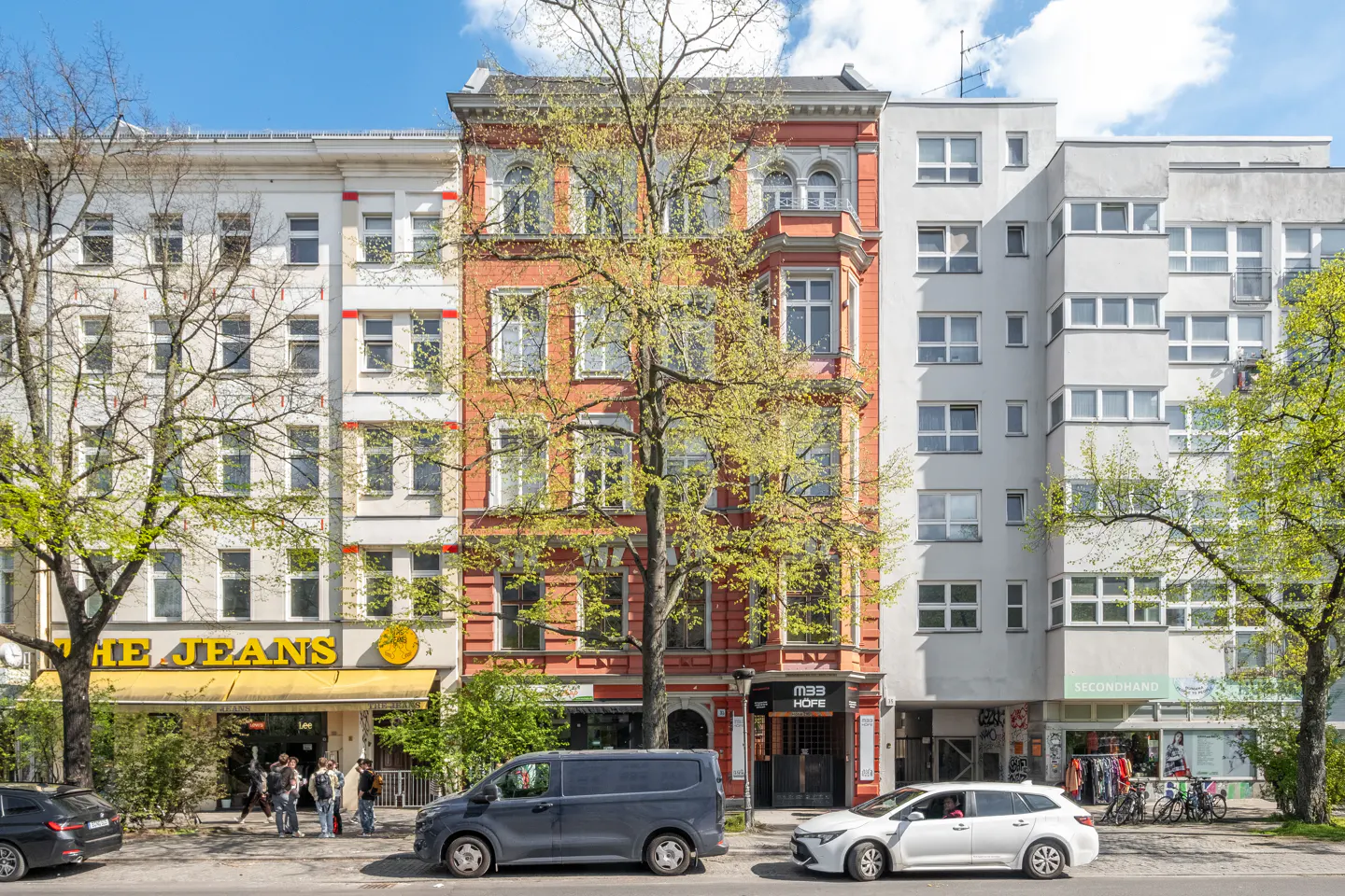 Street view of a red brick building with a tree in front, flanked by white buildings with shops on the ground floor.