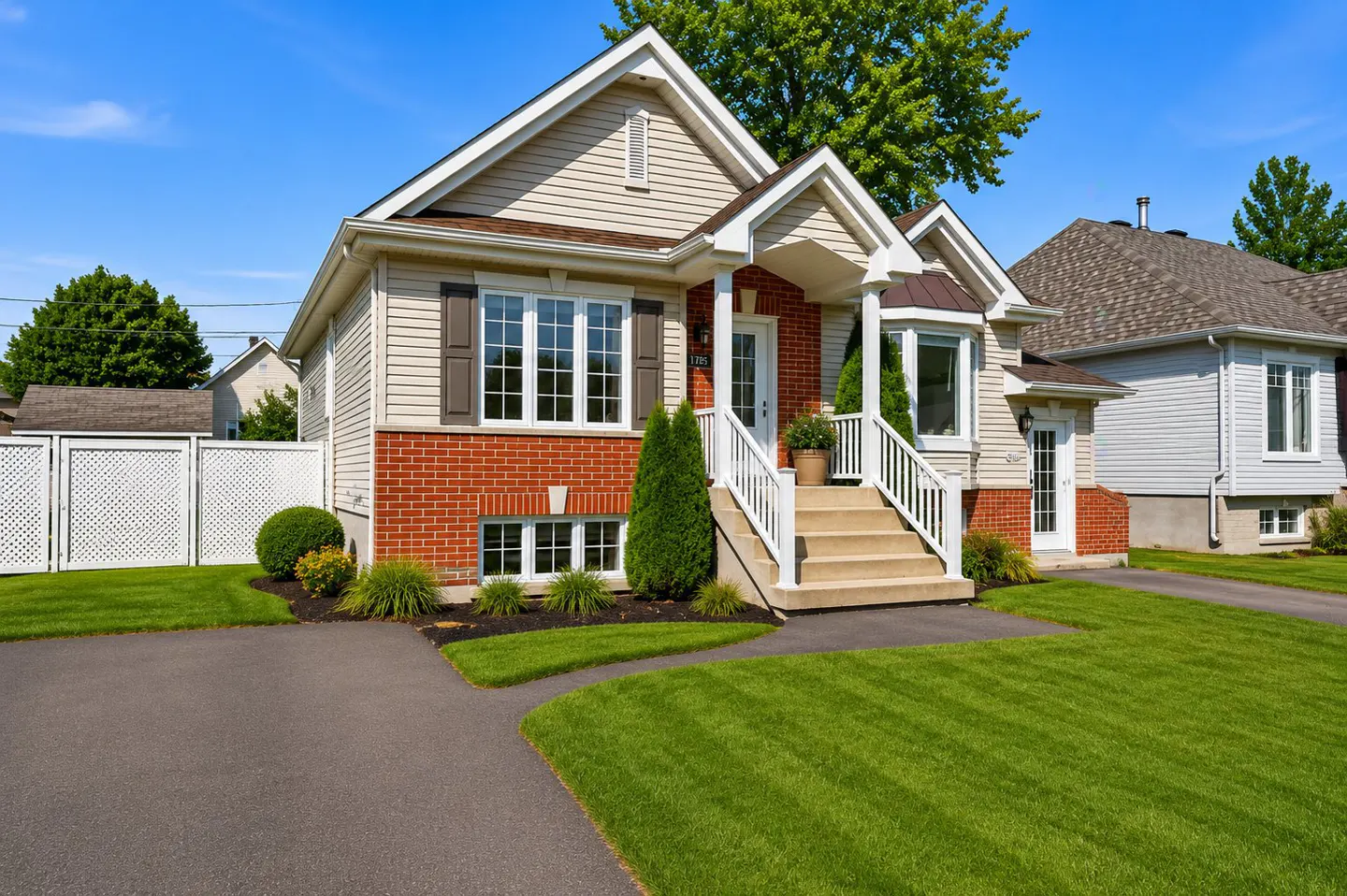 A charming one-story house with beige siding, red brick accents, a white porch, and a well-manicured green lawn.