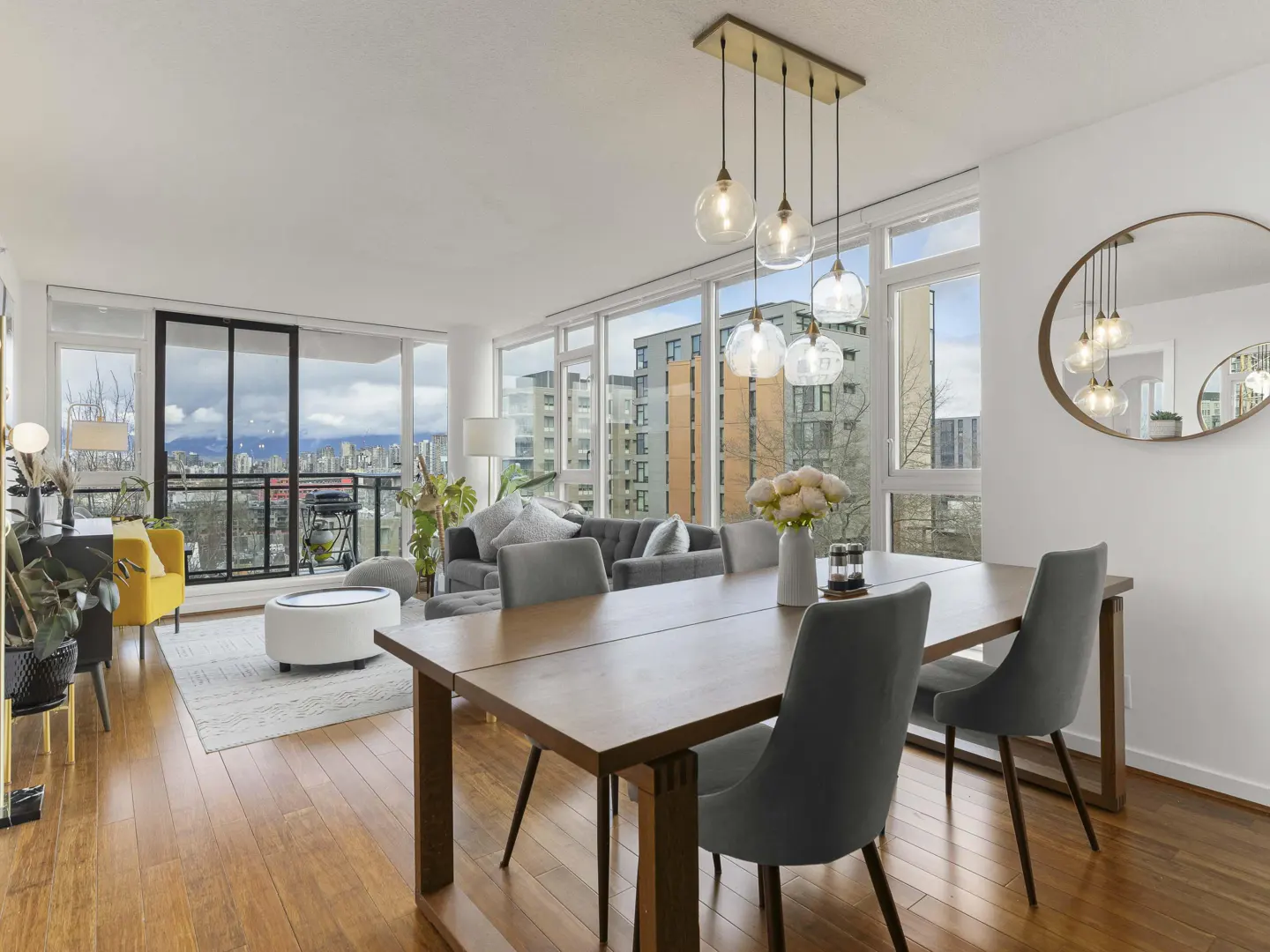 Bright, open-concept condo with wood floors, dining table, and city views. Gray sofa and yellow chair in the living area. Balcony with black railings.