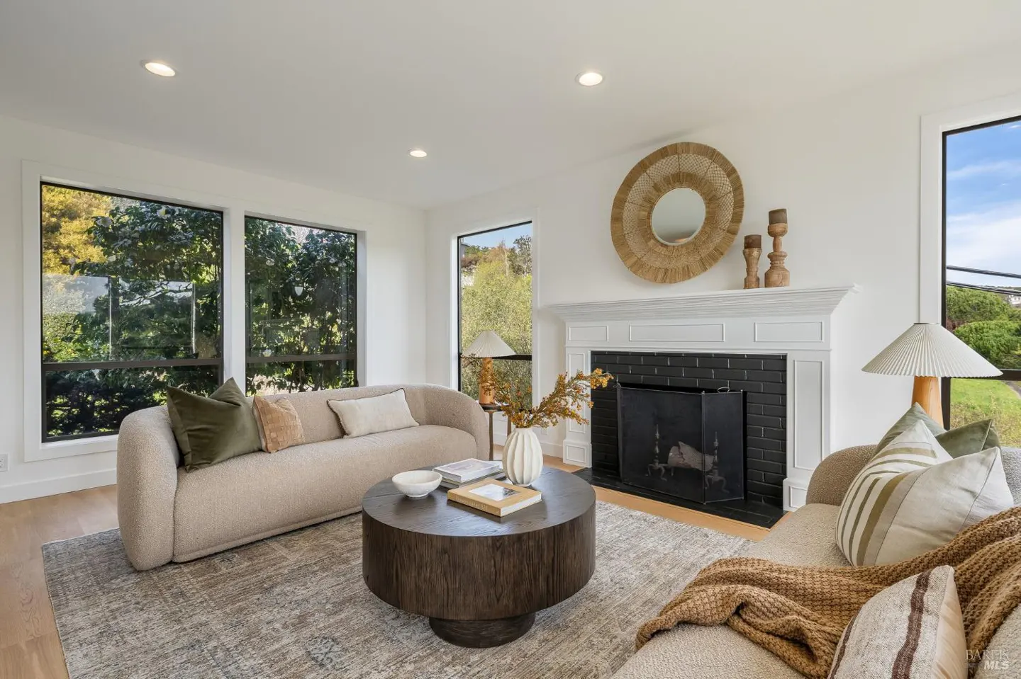 Bright living room with two sofas, a round wood coffee table, and a fireplace with a white mantel and round mirror above.