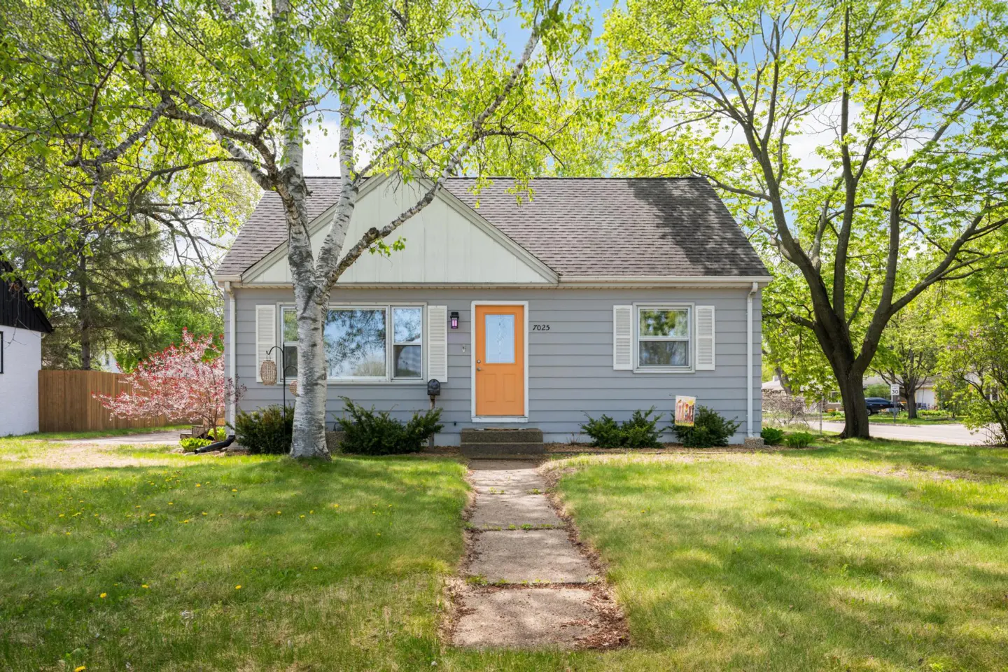 A gray one-story house with an orange door and white trim, surrounded by green trees and a lawn.
