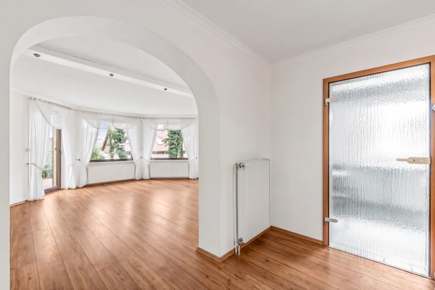 Interior view of a room with wood floors, white walls, and an arched doorway leading to a bright living area with windows and white curtains. A textured glass door is on the right.