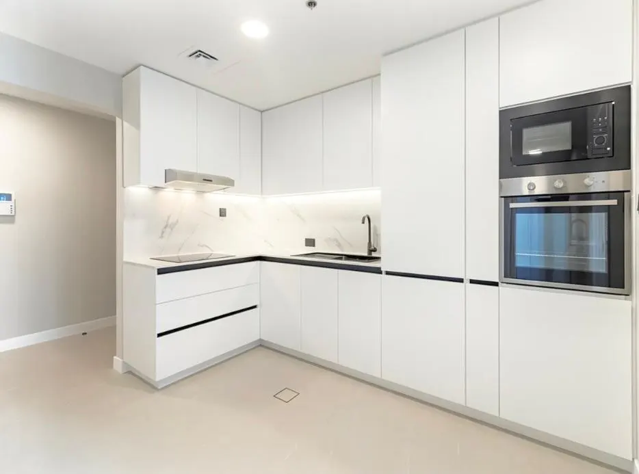 A modern kitchen with white cabinets, black countertops, and stainless steel appliances. A black faucet is visible. The floor is light beige.